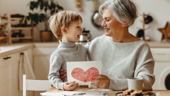 Senior woman at kitchen table holding paper with a heart on it with her grandson